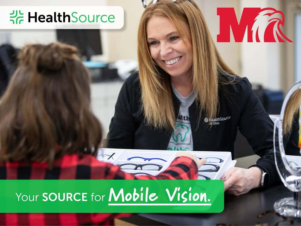 A smiling HealthSource staff member helps a child select eyeglass frames from a tray, with a tabletop mirror nearby. The HealthSource logo appears in the upper left, and Milford’s red “M” and Eagle logo appears in the upper right. A green banner reads, “Your SOURCE for Mobile Vision.”
