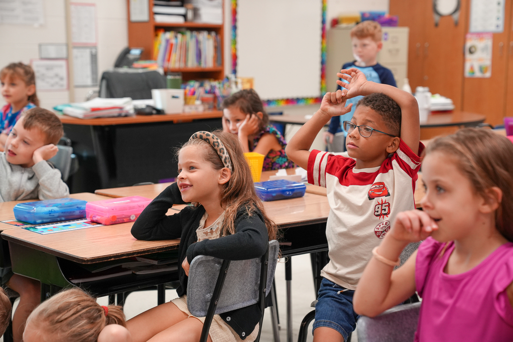 Elementary students listen to a classroom presentation, with one student raising both arms above his head.
