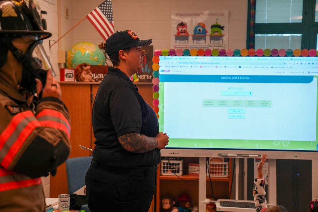 A firefighter in full protective gear stands beside another fire department representative speaking to students in a classroom. A student raises their hand to ask a question.
