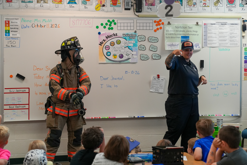 A firefighter wearing full protective gear and a fire department representative speak to a classroom of elementary students about firefighting and safety.
