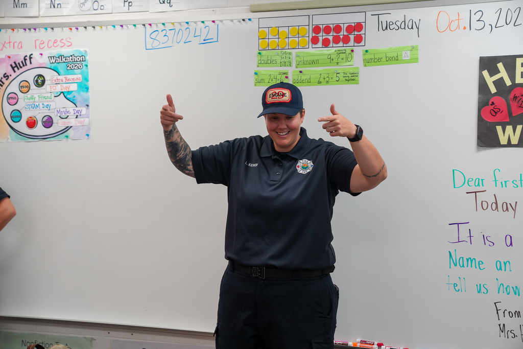 A fire department representative smiles and gestures with both hands while speaking to elementary students in front of a classroom whiteboard.