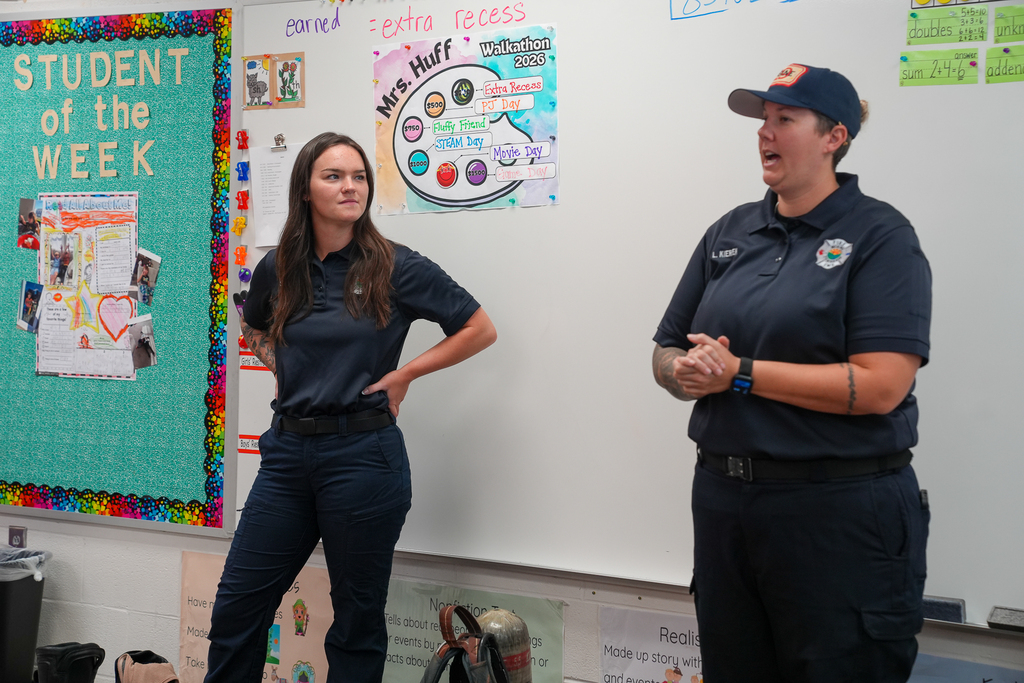 Two fire department representatives speak to elementary students in a classroom, with a student recognition bulletin board and classroom decorations behind them.