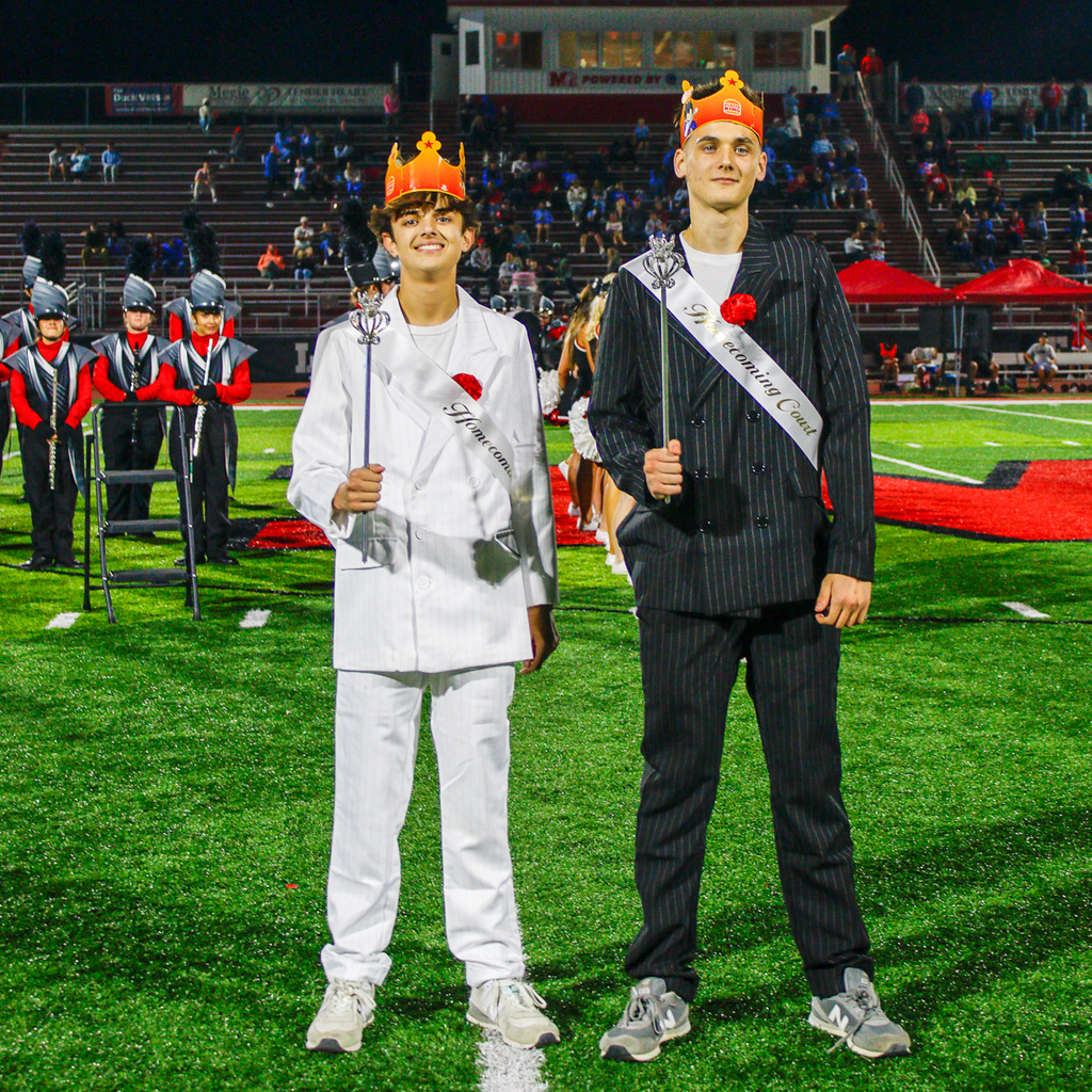 Two students wearing orange crowns and formal suits, one white and one black pinstriped, pose together while holding decorative scepters.