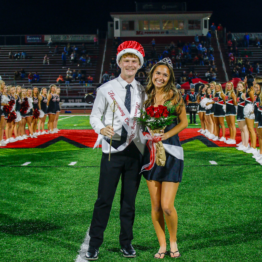 Two students wearing crowns and Homecoming Court sashes pose together. One holds a scepter, and the other holds a bouquet of red roses.