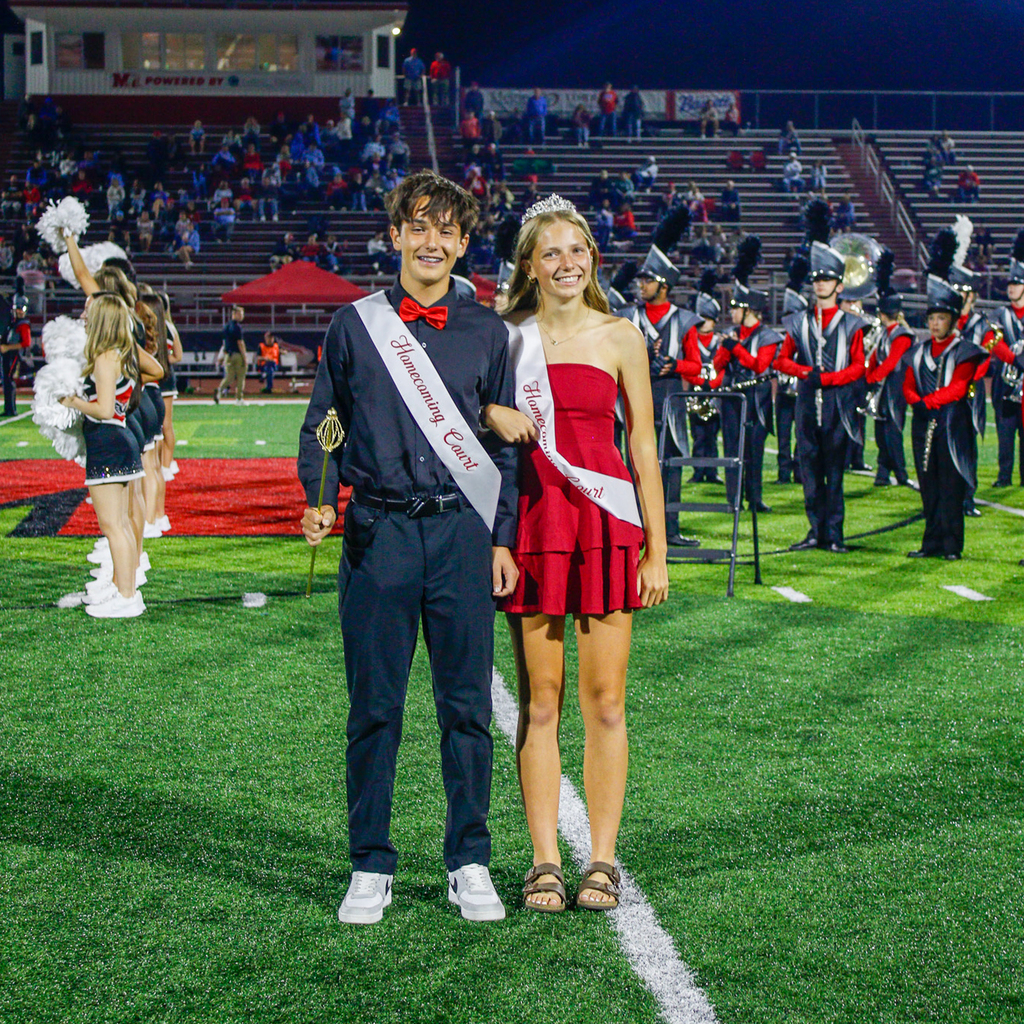 Two Homecoming Court members pose arm in arm. One wears a black outfit with a red bow tie, while the other wears a red dress and tiara.