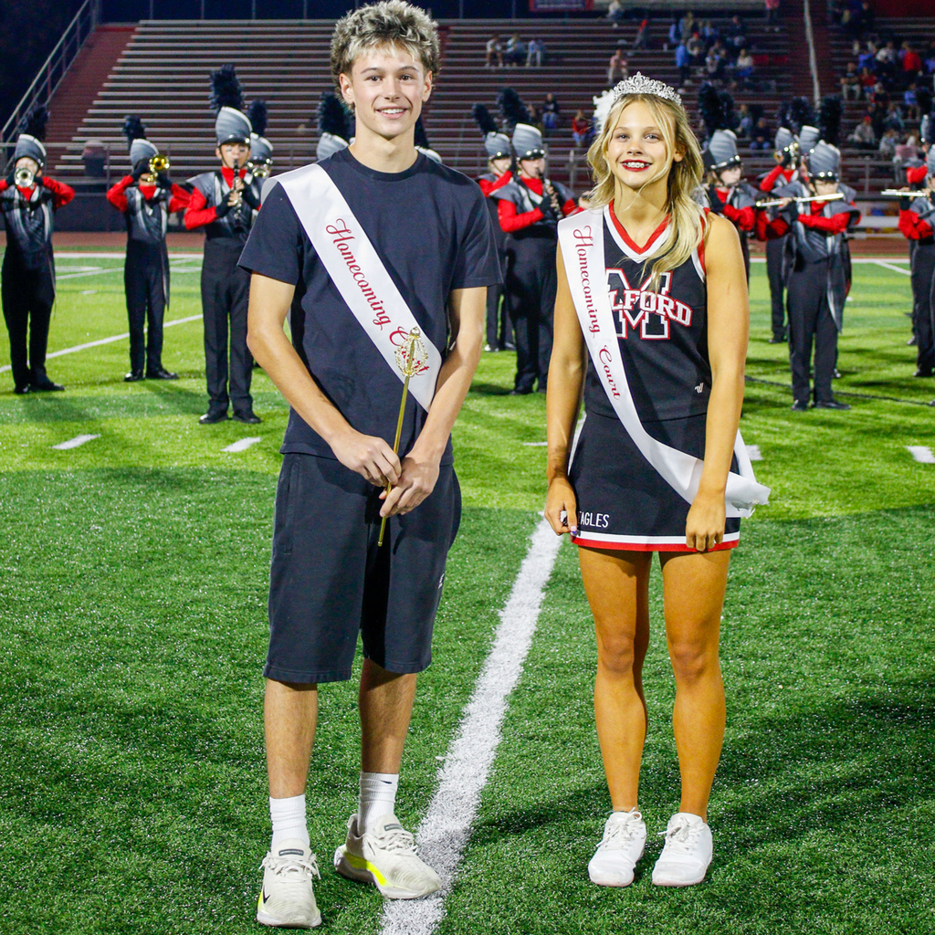 Two students pose in front of the marching band. One wears a navy shirt and shorts, while the other wears a Milford cheerleading uniform and tiara.