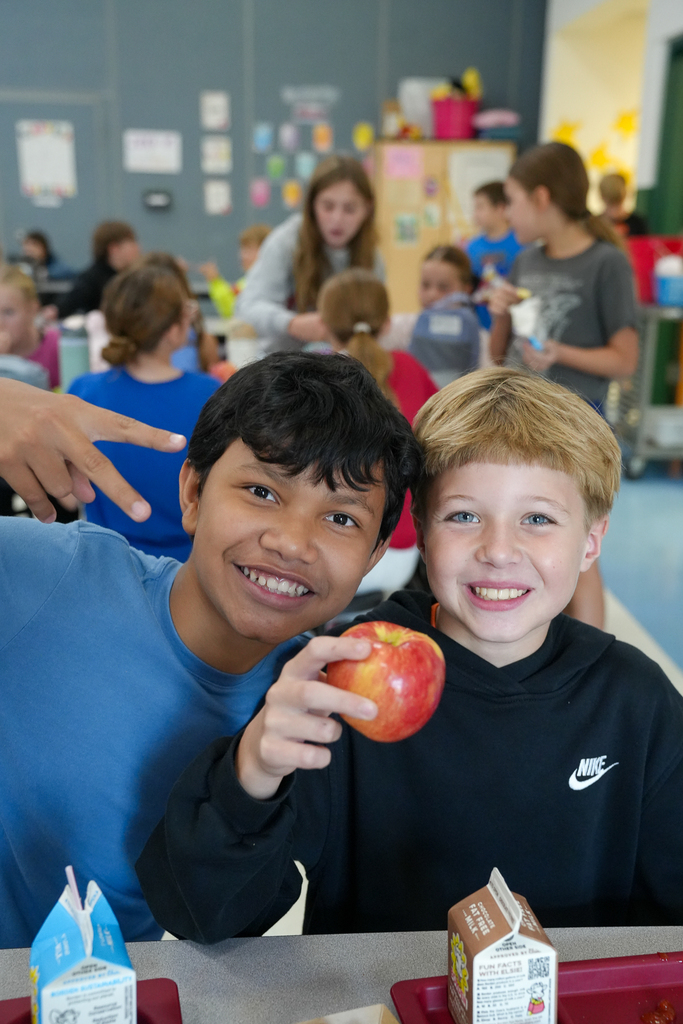 Two students lean together and smile at the camera. One holds up a red apple while the other makes a peace sign.