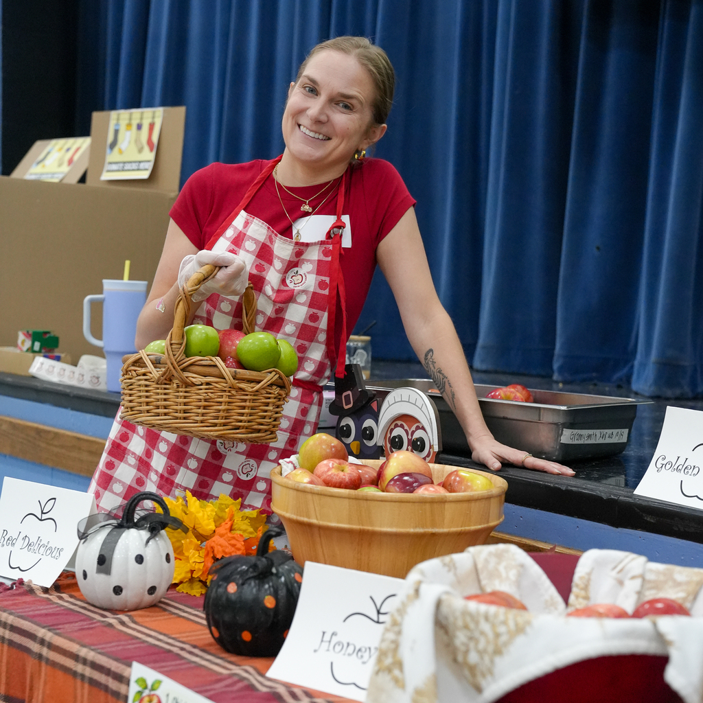 A smiling School Nutrition team member holds a basket of red and green apples behind a display decorated with pumpkins, fall leaves and signs identifying apple varieties.