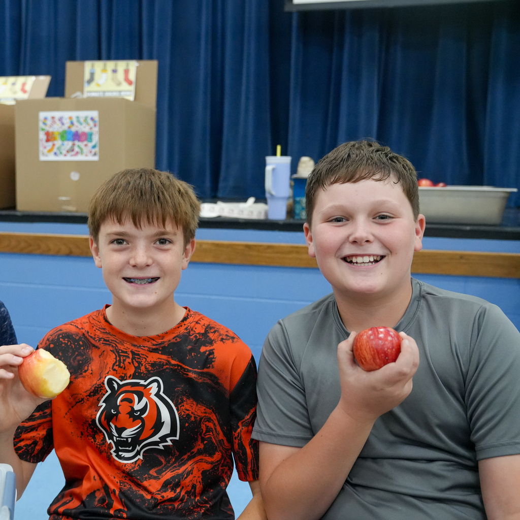 Three students smile and hold up red and green apples while seated together at a cafeteria table.