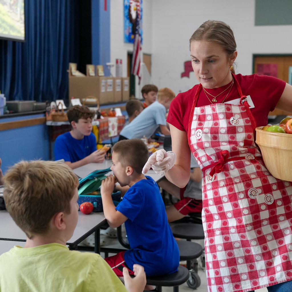 A School Nutrition team member wearing an apple-patterned apron carries a wooden bowl of apples and talks with a student in the cafeteria.