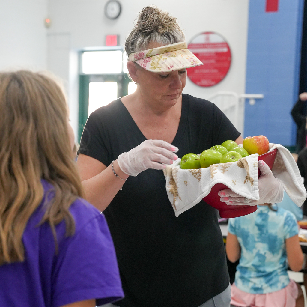 A School Nutrition team member wearing gloves and a floral visor holds a bowl of green and red apples beside a student.