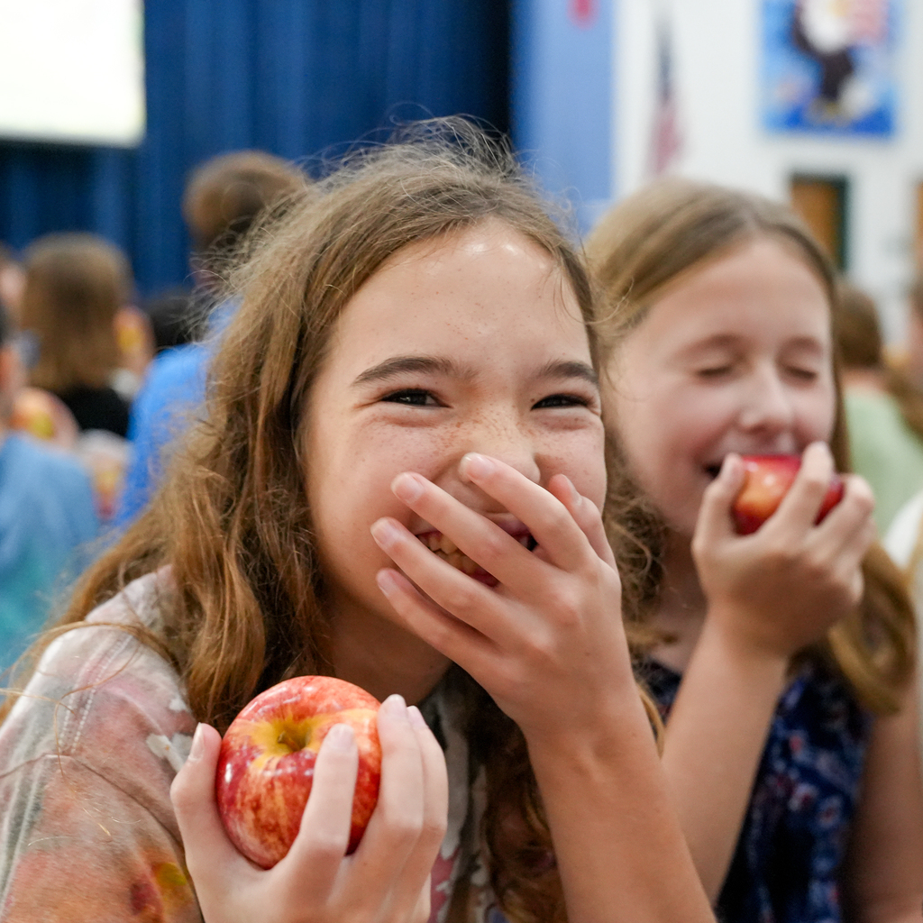 Two students enjoy red apples at a cafeteria table, with one taking a bite and the other smiling.