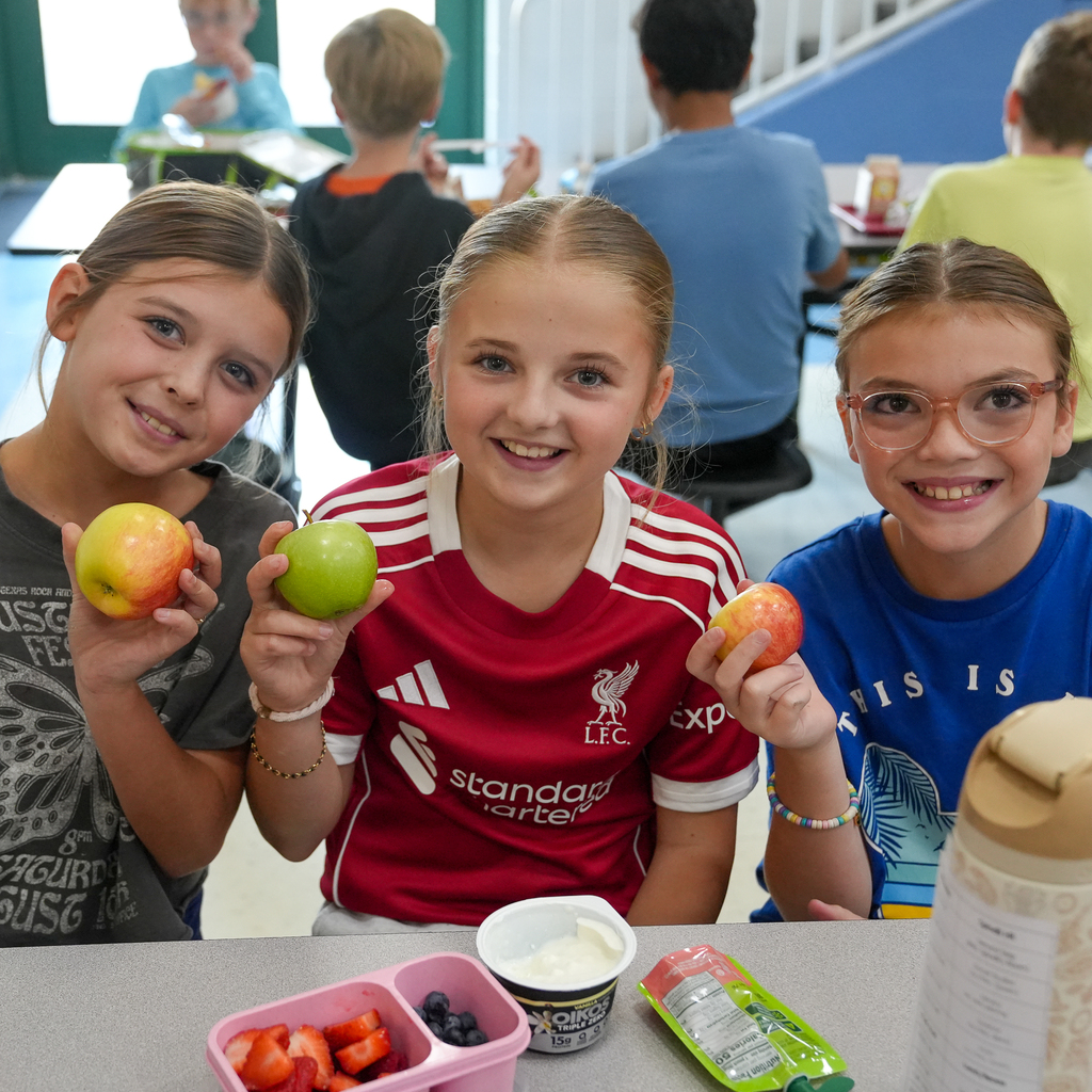 Three students smile and hold up red and green apples while seated together at a cafeteria table.