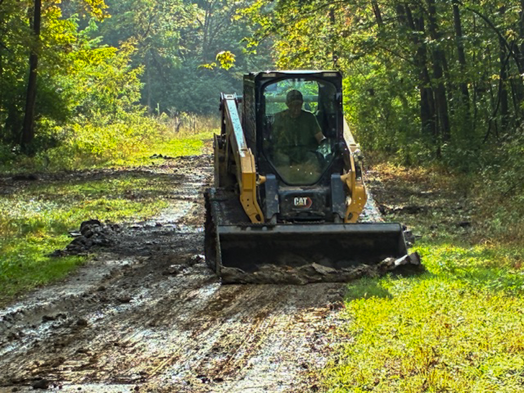 A student operates a yellow compact track loader to level a muddy section of the cross-country course, surrounded by trees.