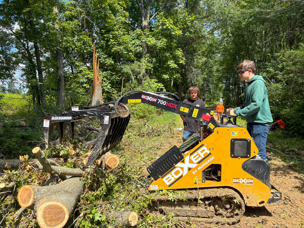 A student operates a compact loader with a grapple attachment to move cut tree limbs beside the wooded cross-country course as another student looks on.