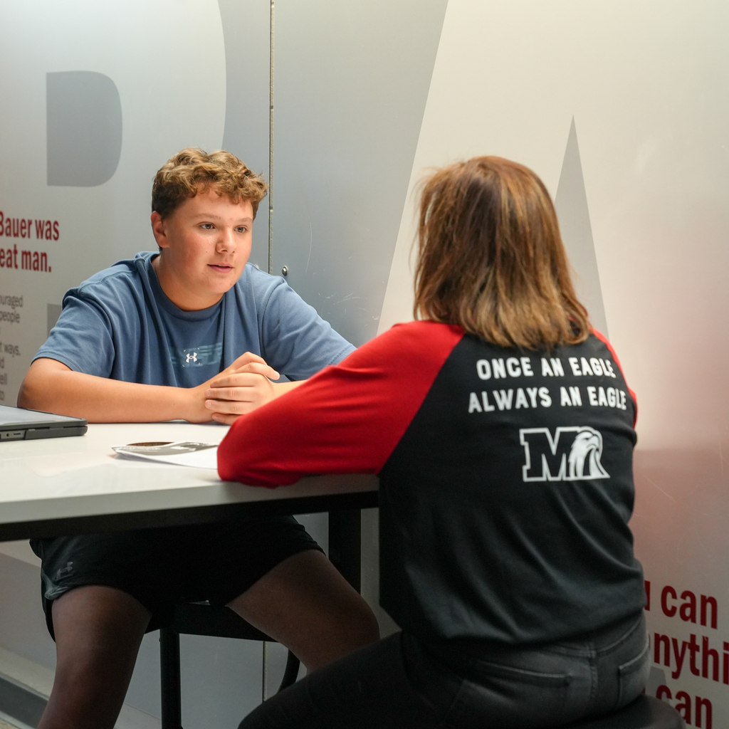 A student in a blue shirt sits with hands clasped while speaking with an interviewer whose Milford shirt reads, “Once an Eagle, Always an Eagle.”
