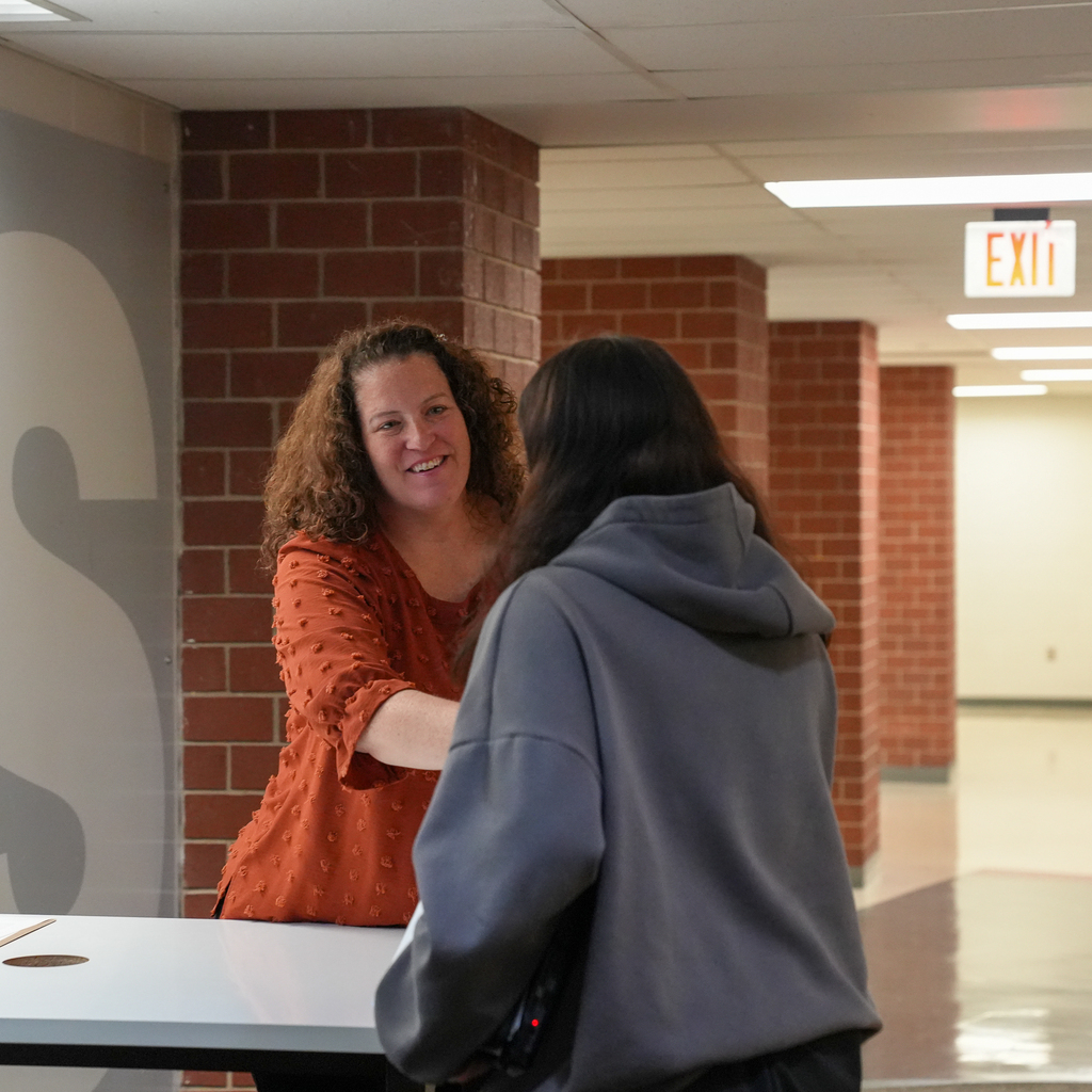 An adult interviewer in an orange blouse smiles while greeting a student wearing a gray hoodie across a table.