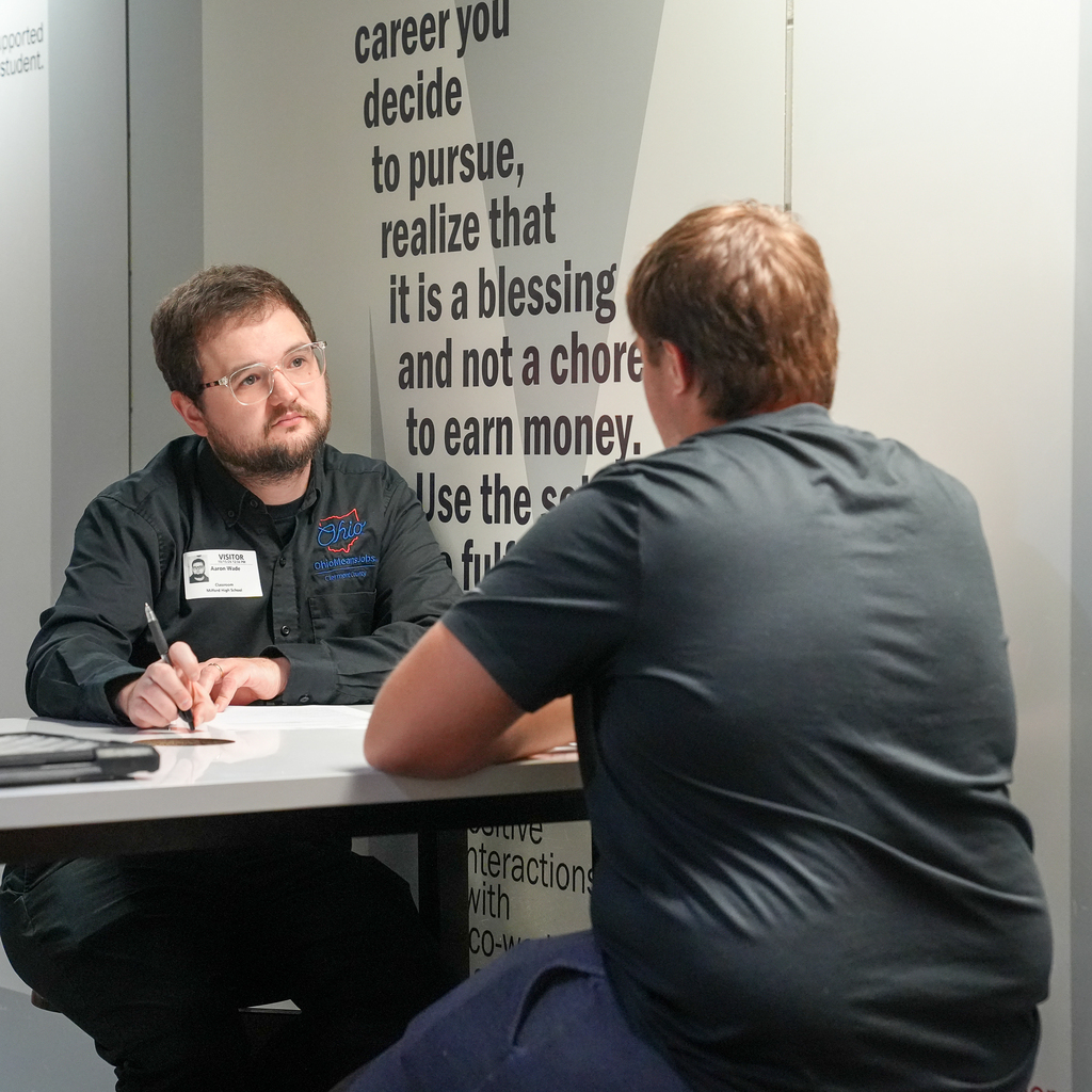 An interviewer wearing an OhioMeansJobs shirt listens to a student across a table, holding a pen over interview paperwork.