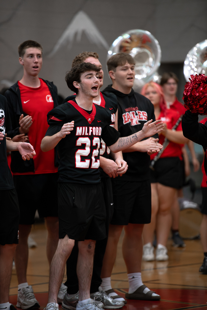 Milford High School football players and students cheer together in the gym. A student wearing a black Milford football jersey with the number 22 stands in the foreground with his arms extended.