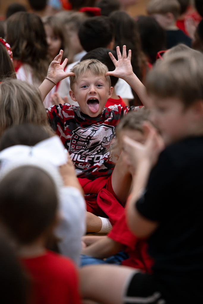 An elementary student wearing a red, black and white Milford shirt makes a silly face with his tongue out and both hands raised above his head while sitting among other students.
