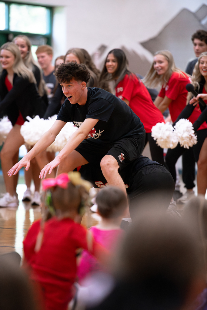 A Milford High School student in a black T-shirt and shorts leans forward with his arms extended during a school spirit activity. Cheerleaders and other students gather behind him in the gym.