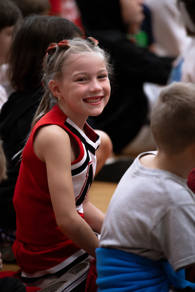 : A young Milford cheerleader in a red, white and black uniform smiles toward the camera while sitting among students during a school spirit event.