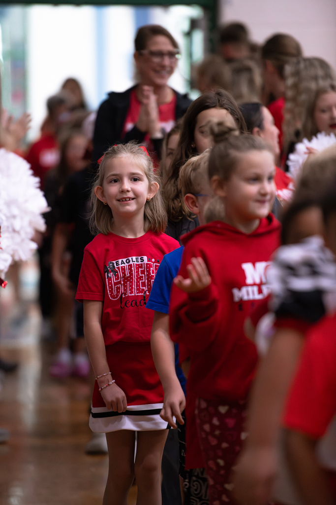 An elementary student wearing a red Eagles Cheer T-shirt and matching cheer skirt walks through a crowd of students and staff, with cheerleaders holding pom-poms along the sides.