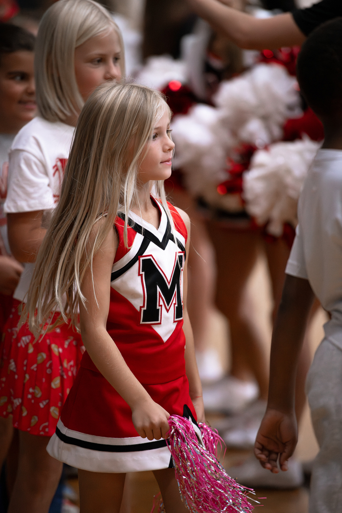 An elementary student wearing a red Eagles Cheer T-shirt and matching cheer skirt walks through a crowd of students and staff, with cheerleaders holding pom-poms along the sides.