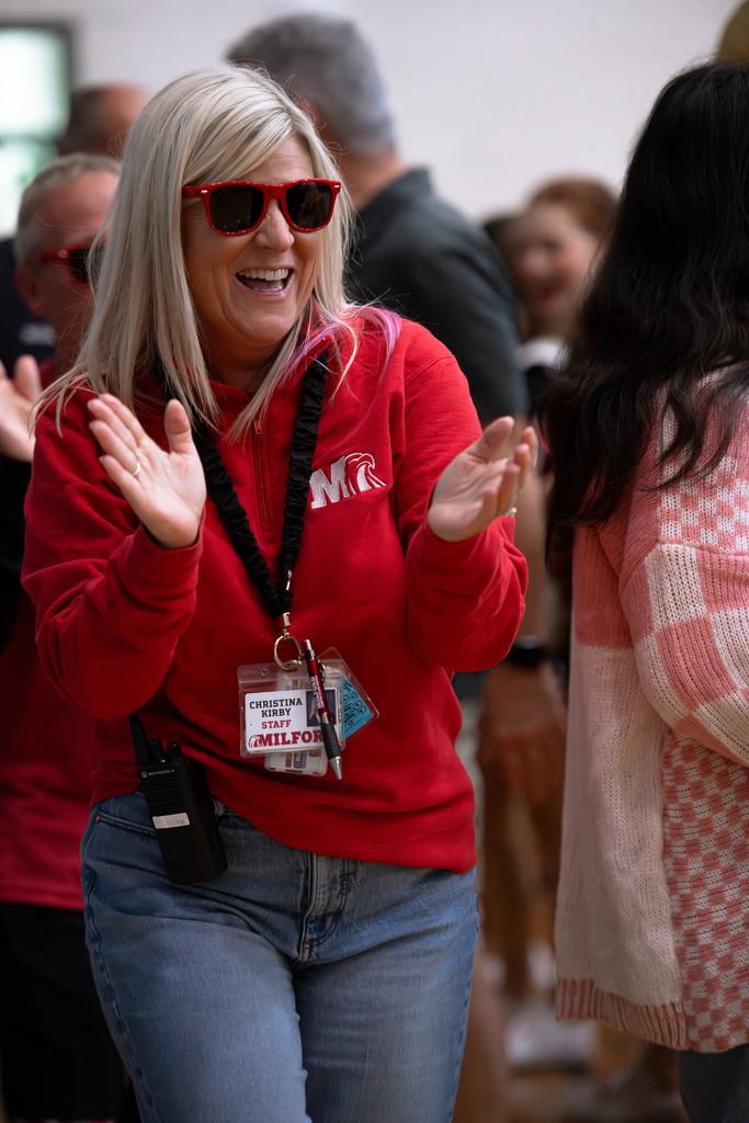 A Milford staff member wearing a red Milford sweatshirt and red sunglasses smiles and claps during a school spirit event.