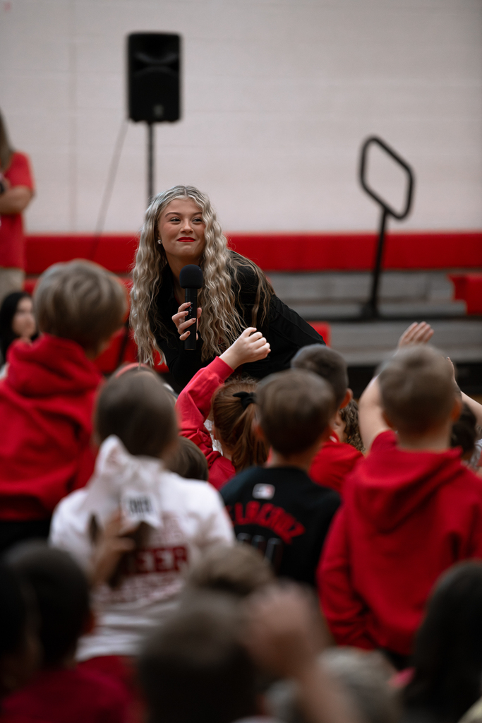 A Milford High School student with long blonde hair holds a microphone and speaks with elementary students during a school spirit event in the gymnasium. Several students wearing red and black raise their hands to participate.