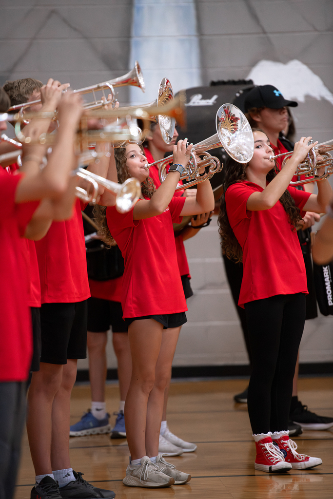 Milford High School marching band members wearing red shirts play brass instruments inside the gymnasium, with percussionists visible behind them.