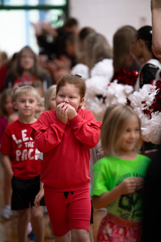 An elementary student dressed in red covers her mouth with both hands while walking through a crowd of students and cheerleaders holding red and white pom-poms.