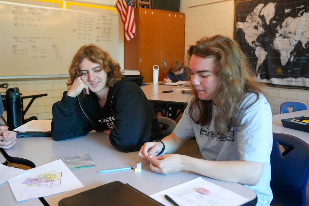 A student assembles small pastel bricks while a classmate watches. A colored drawing of a brick structure lies on the table nearby.