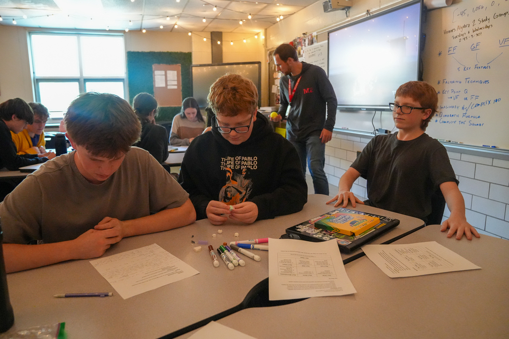 Three students sit at grouped desks as the center student assembles small bricks. A teacher stands behind them while other groups work nearby.