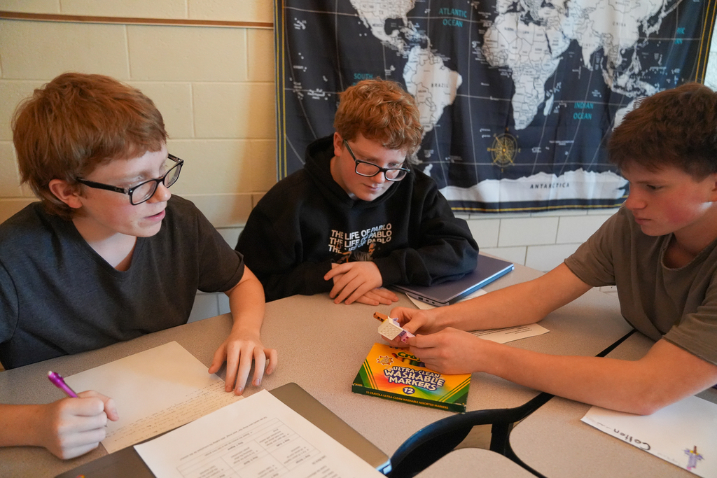 Three students work together at a table. One writes notes while another holds a small brick model for the group to examine.  AP
