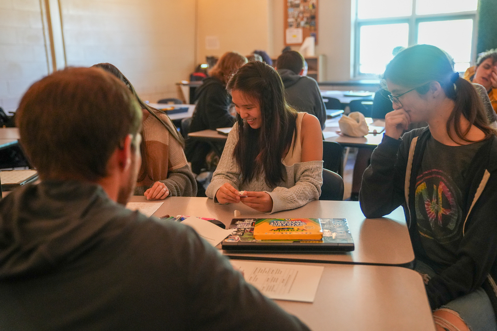 A student smiles while holding small building pieces as classmates gather around the table during a classroom activity.