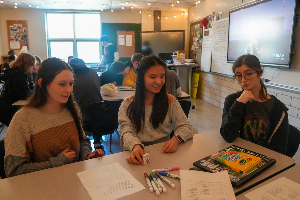 Three students sit around a classroom table with a small pastel brick model, worksheets and markers in front of them.