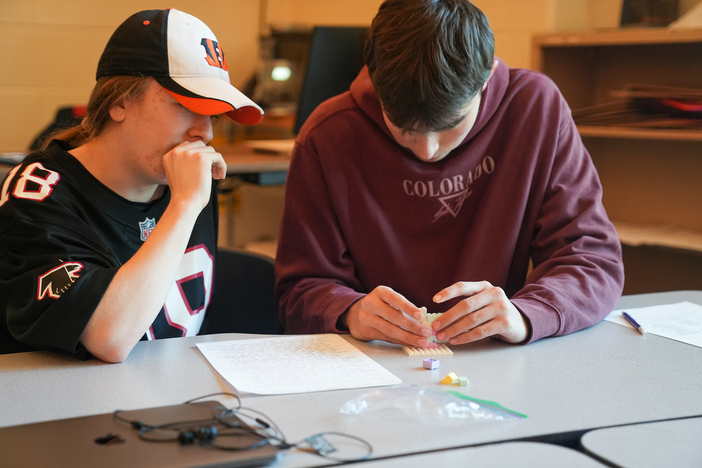 Two students examine a brick structure as one carefully adds pieces to it. Handwritten notes and extra bricks sit beside them.
