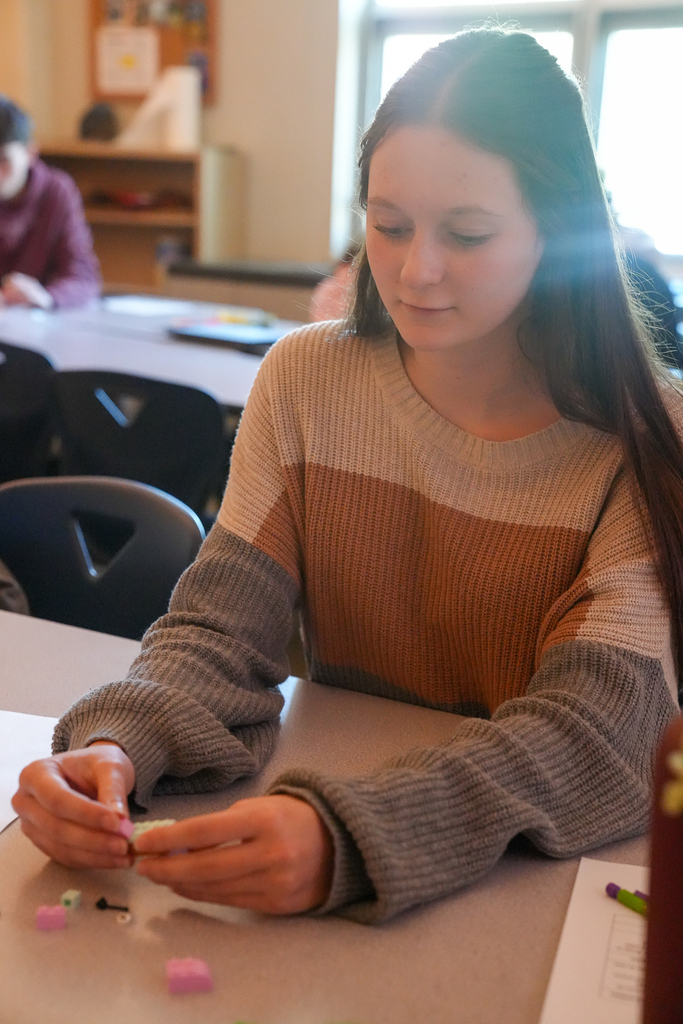 A student in a striped sweater connects pastel bricks at a classroom desk, with loose pieces spread across the tabletop.
