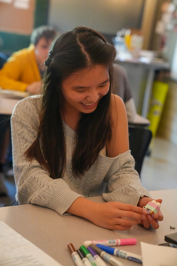 A student smiles while holding a small model made from colorful interlocking bricks, with markers and papers on the classroom desk.