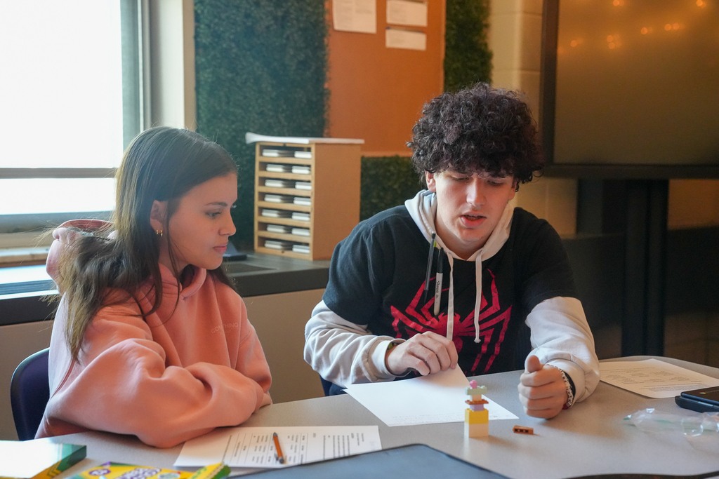Two students look at a tall, colorful brick model on their table. One gestures beside the model, with worksheets spread nearby.