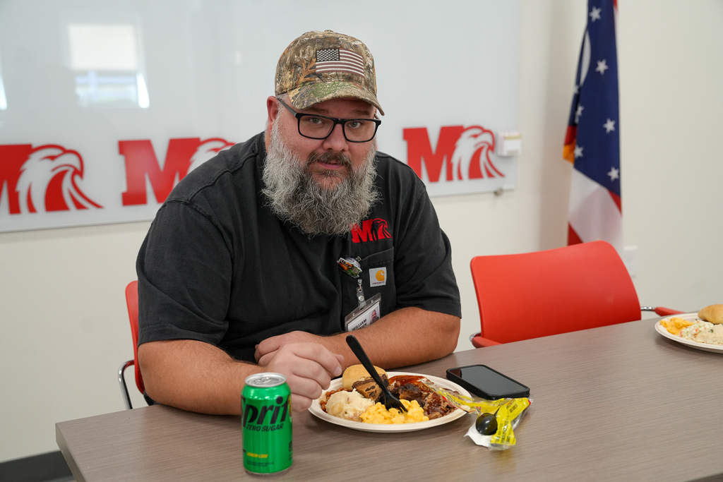 A Milford Schools custodial staff member with a gray beard, camouflage baseball cap and black Milford shirt sits at a table with a plate of food and a green soda can during an appreciation luncheon.