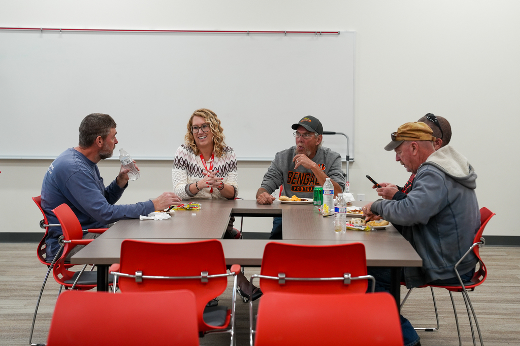 Milford Schools custodial staff members gather around tables during an appreciation luncheon. Several staff members talk while enjoying their meals, with red Milford logos displayed on the wall behind them.