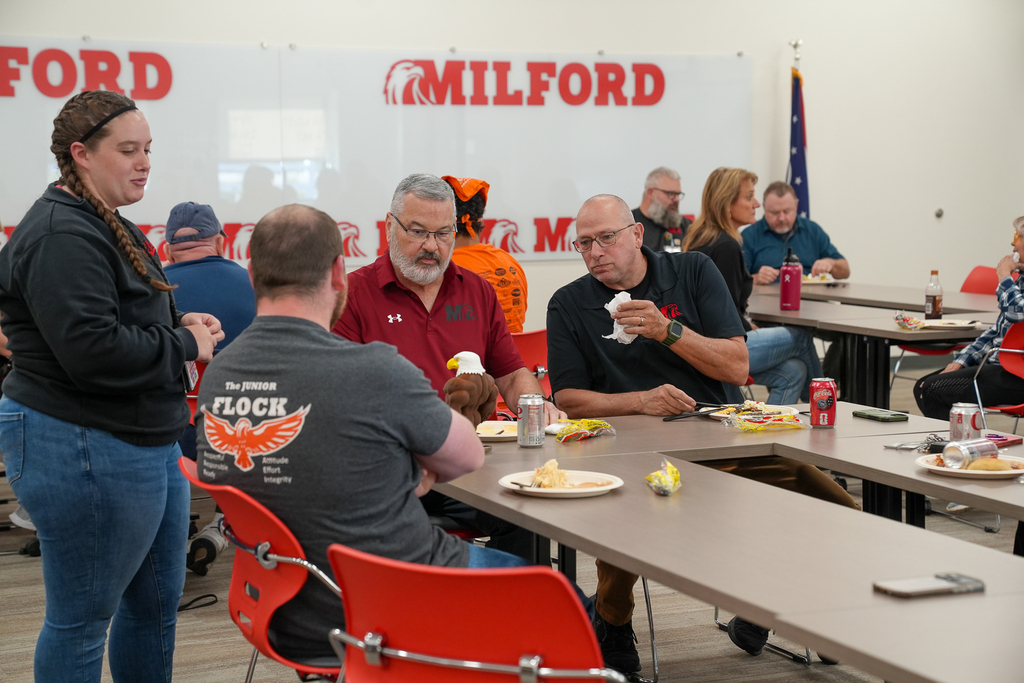 Four Milford Schools staff members sit around a table eating lunch and talking. Red chairs, plates of food and a whiteboard are visible in the meeting room.