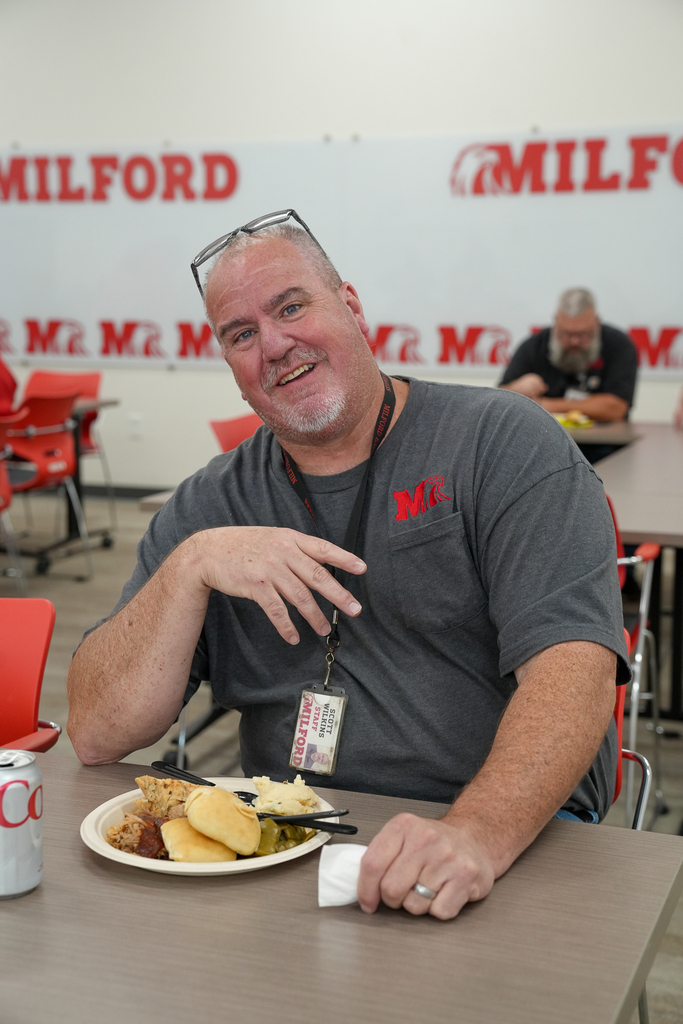 A smiling Milford Schools custodial staff member wearing a gray Milford shirt and staff ID badge sits at a table with a plate of food during an appreciation luncheon.