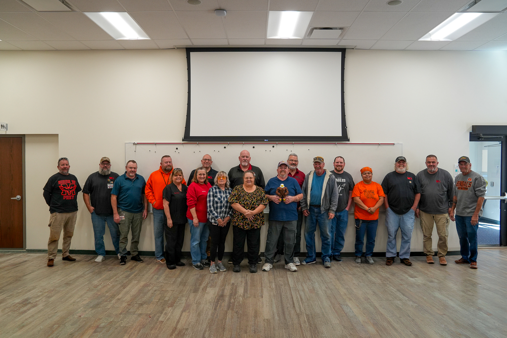 Milford Schools custodial staff members pose together for a group photo during their appreciation luncheon. The group stands in front of a whiteboard beneath a large projection screen.