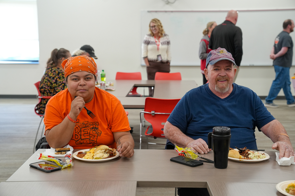 Two Milford Schools custodial staff members sit together at a table enjoying an appreciation luncheon. One wears an orange shirt and matching bandana, while the other wears a blue shirt and baseball cap. Plates of food sit in front of them.