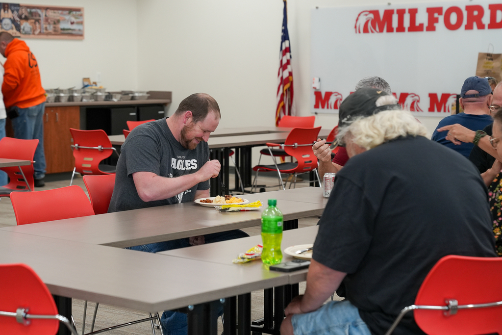 Milford Schools custodial staff members enjoy lunch and conversation around tables with red chairs. A staff member wearing a gray Milford Eagles shirt smiles while eating, with Milford signage visible on the wall.