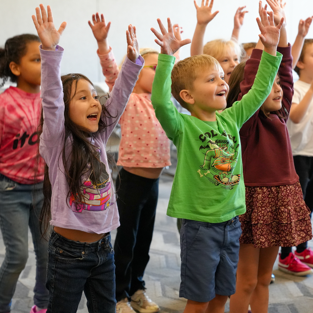 Mulberry kindergarten students smile and raise both hands overhead during a group library activity.