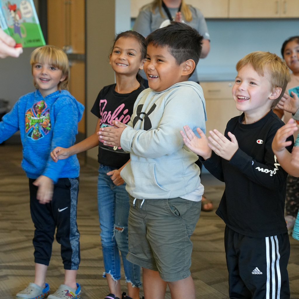 Students smile and clap while looking toward an adult holding a picture book.